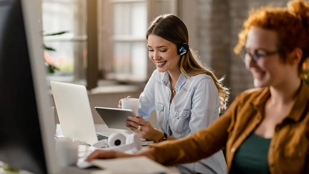 Duas atendentes sorrindo enquanto conversam com clientes pelo computador.
